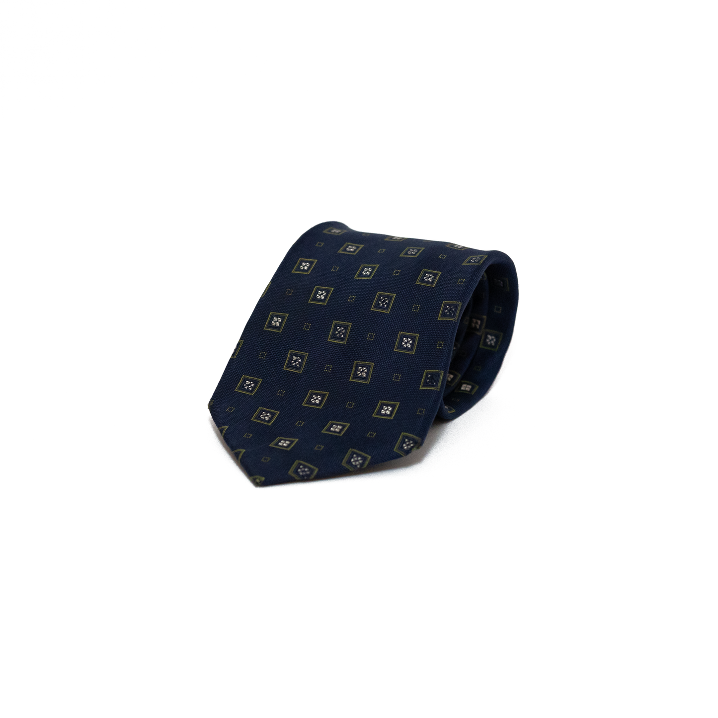 Vintage silkeslips fra Sartoria Italiana i dyp marineblå, dekorert med små rombemotiv i gylne og olivengrønne toner spredt jevnt over hele lengden. Den fine vevingen i silken gir overflaten dybde og et subtilt glans, og det rikt detaljerte mønsteret skaper et klassisk italiensk uttrykk.