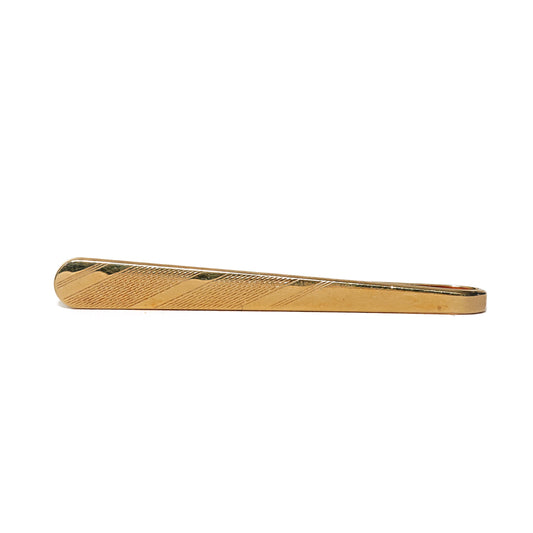 Vintage slipsnål fra 70-tallet i avlang form og gulltone. Et midtfelt med fine, diagonale linjer brytes opp av polerte striper som skjærer over mønsteret, og kontrasten mellom det matte og det blanke gir et grafisk og dynamisk uttrykk. Den slanke formen og de rene detaljene gir slipsnålen et moderne preg. Et stilrent valg for mannen som setter pris på diskrete linjer og tidens designspråk.