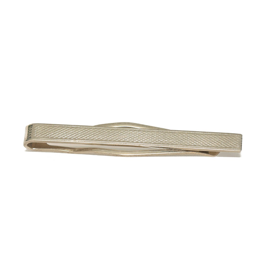 Vintage slipsnål fra 60-tallet i avlang form og sølvtone, dekorert med et fint hakket rutemønster som dekker hele lengden. Den tette teksturen fanger lyset i et dempet, men levende spill og gir slipsnålen et raffinert preg uten å bli prangende. Et tidløst valg for mannen som setter pris på diskrete detaljer og klassisk håndverk.