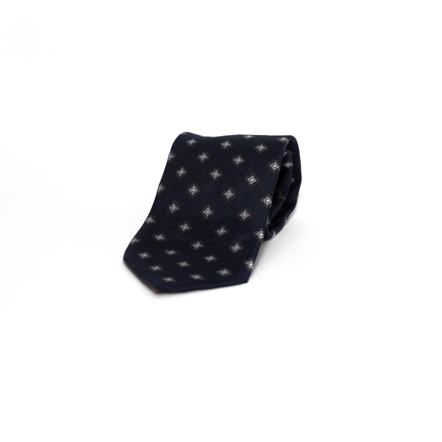 Vintage silkeslips fra Andrew's Ties i dyp marineblå, dekorert med små geometriske rombemotiv i lyseblå og brune toner spredt jevnt over hele lengden. Den fine diagonale vevingen i silken gir overflaten et subtilt glans, og de dempede fargene i mønsteret skaper et raffinert og diskret uttrykk.