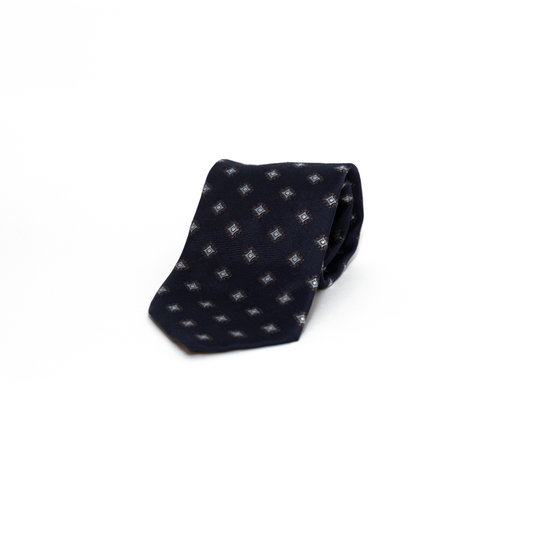 Vintage silkeslips fra Andrew's Ties i dyp marineblå, dekorert med små geometriske rombemotiv i lyseblå og brune toner spredt jevnt over hele lengden. Den fine diagonale vevingen i silken gir overflaten et subtilt glans, og de dempede fargene i mønsteret skaper et raffinert og diskret uttrykk.