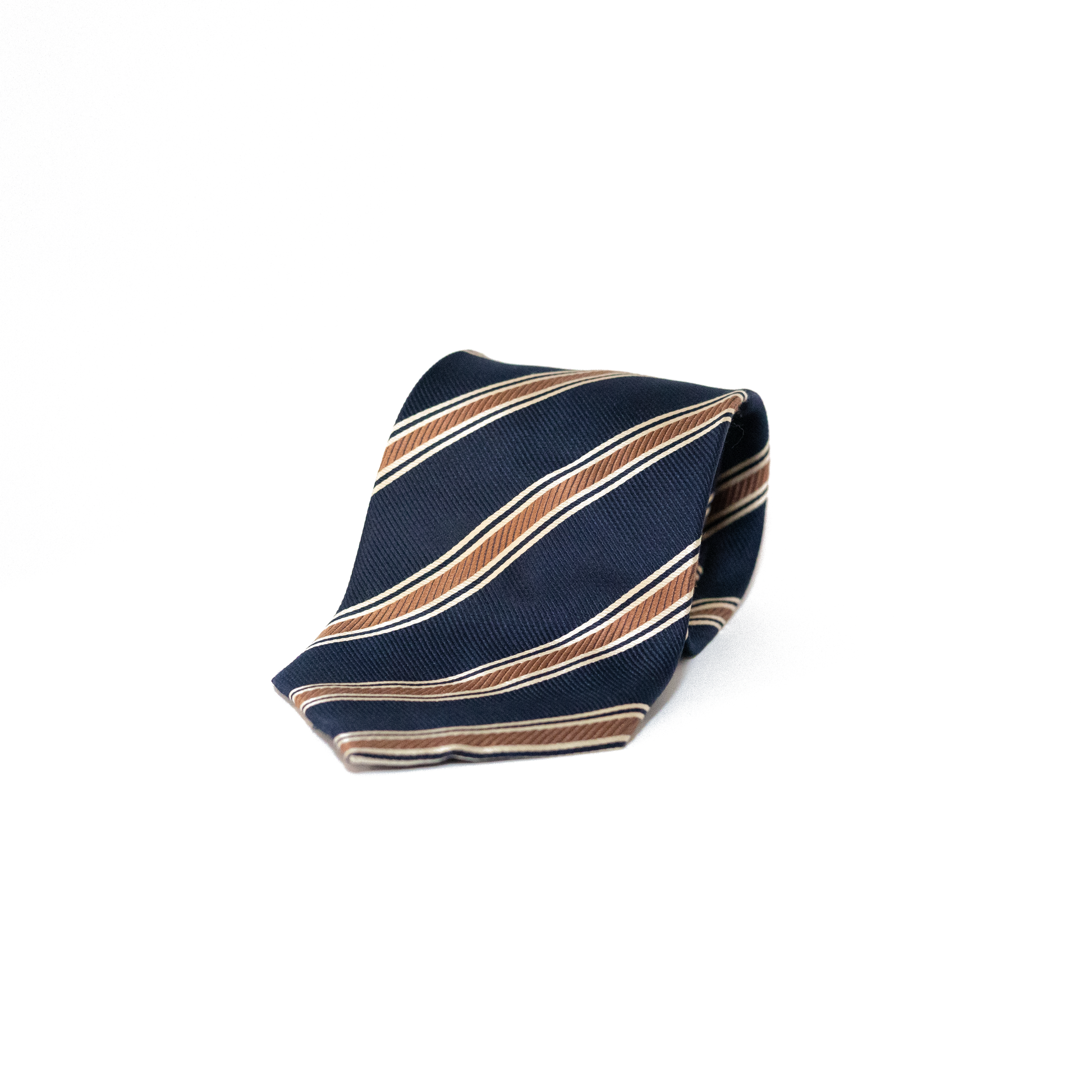 Mørkeblått vintage slips fra Monsieur Gaspar Dido, med diagonale striper i kobberbrunt og tynnere striper i hvitt. Det klassiske stripemønsteret gir slipset et tradisjonelt regimentalt uttrykk, samtidig som det holder seg innenfor et dempet og tidløst utseende.