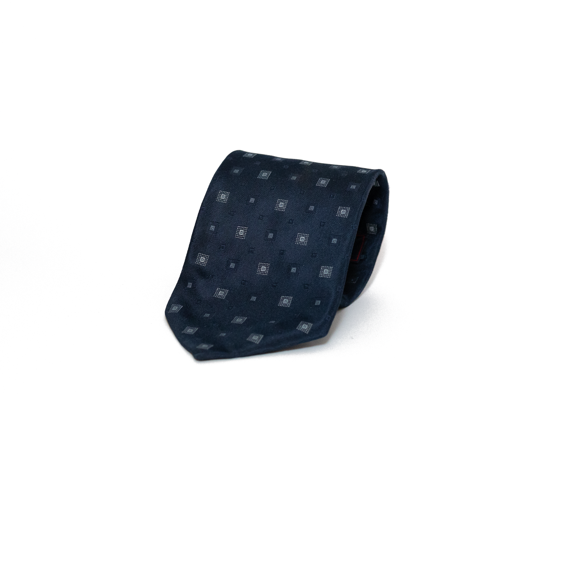 Mørkeblått vintage slips fra italienske Viafrattina, med et diskret geometrisk mønster i små firkanter i sølvtone. Mønsteret gir slipset dybde og variasjon, samtidig som det holder seg innenfor et klassisk og dempet uttrykk.